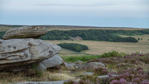 A view from a gritstone ridge, over heather moorland towards a grand building in the distance.